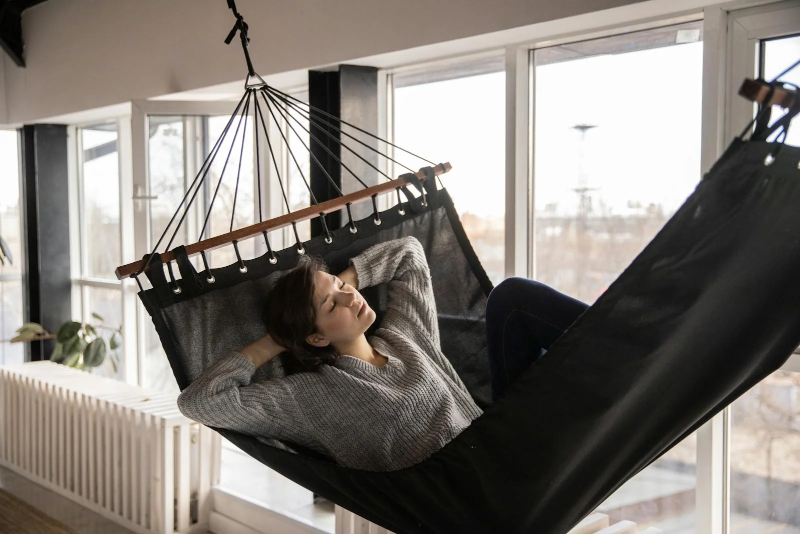 Young lady in casual wear lying in comfortable hammock leaned on hands near window and radiator in apartment in afternoon